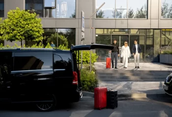 Three adults outside a glass-fronted building near a black van with luggage