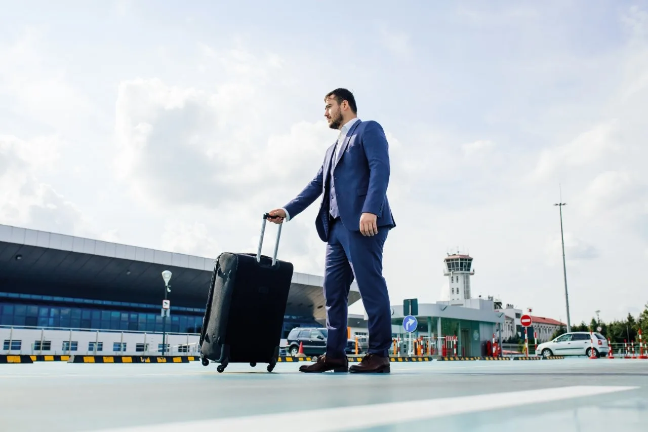 A suited traveler pulling a black suitcase along the exterior of an airport terminal
