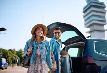 Two travelers with luggage beside a car at a generic airport