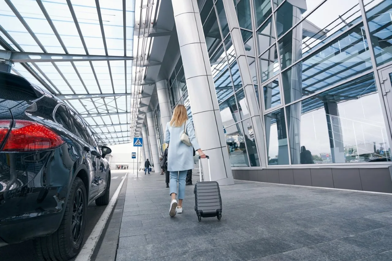 A woman in a light blue suit pulling a gray suitcase beside a black car and glass terminal