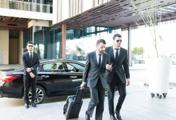 Three men in dark suits beside a black sedan, one pulling a suitcase