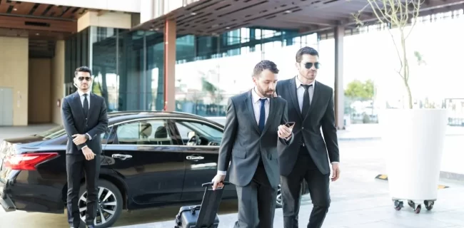 Three men in dark suits beside a black sedan, one pulling a suitcase