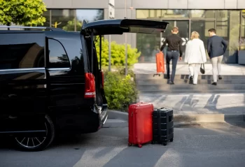 Travelers walking toward glass doors with rolling luggage beside a dark van