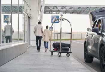 Two travelers walking beside a terminal with a loaded luggage trolley and a dark SUV nearby