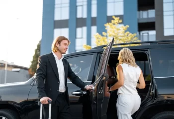 A man holding a vehicle door while a woman enters a black vehicle outside a modern building