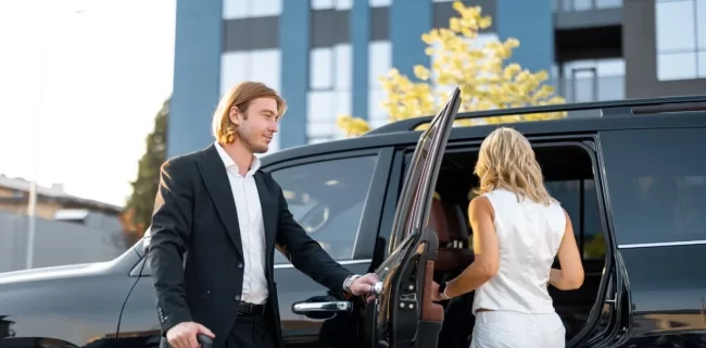 A man holding a vehicle door while a woman enters a black vehicle outside a modern building