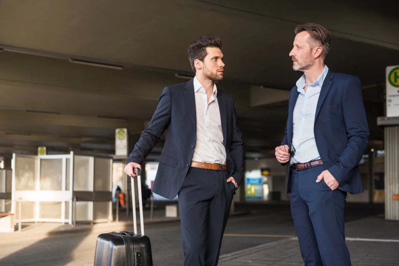 Two men in suits talking beneath a covered exterior area with a suitcase beside them
