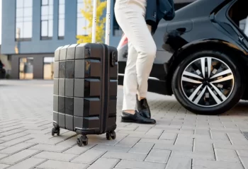 A rolling suitcase beside a traveler in light trousers and a black car outside a building
