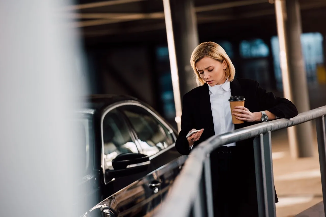 A woman holding a phone and coffee beside a black car and railings
