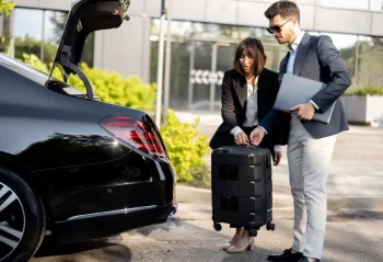 Two colleagues with a suitcase and laptop beside the open trunk of a black sedan