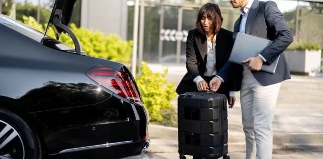 Two colleagues with a suitcase and laptop beside the open trunk of a black sedan