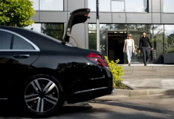 Two adults leaving a glass-fronted building toward a black sedan with its trunk open