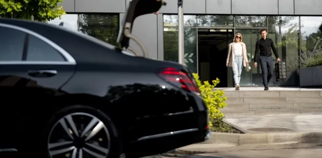 Two adults leaving a glass-fronted building toward a black sedan with its trunk open